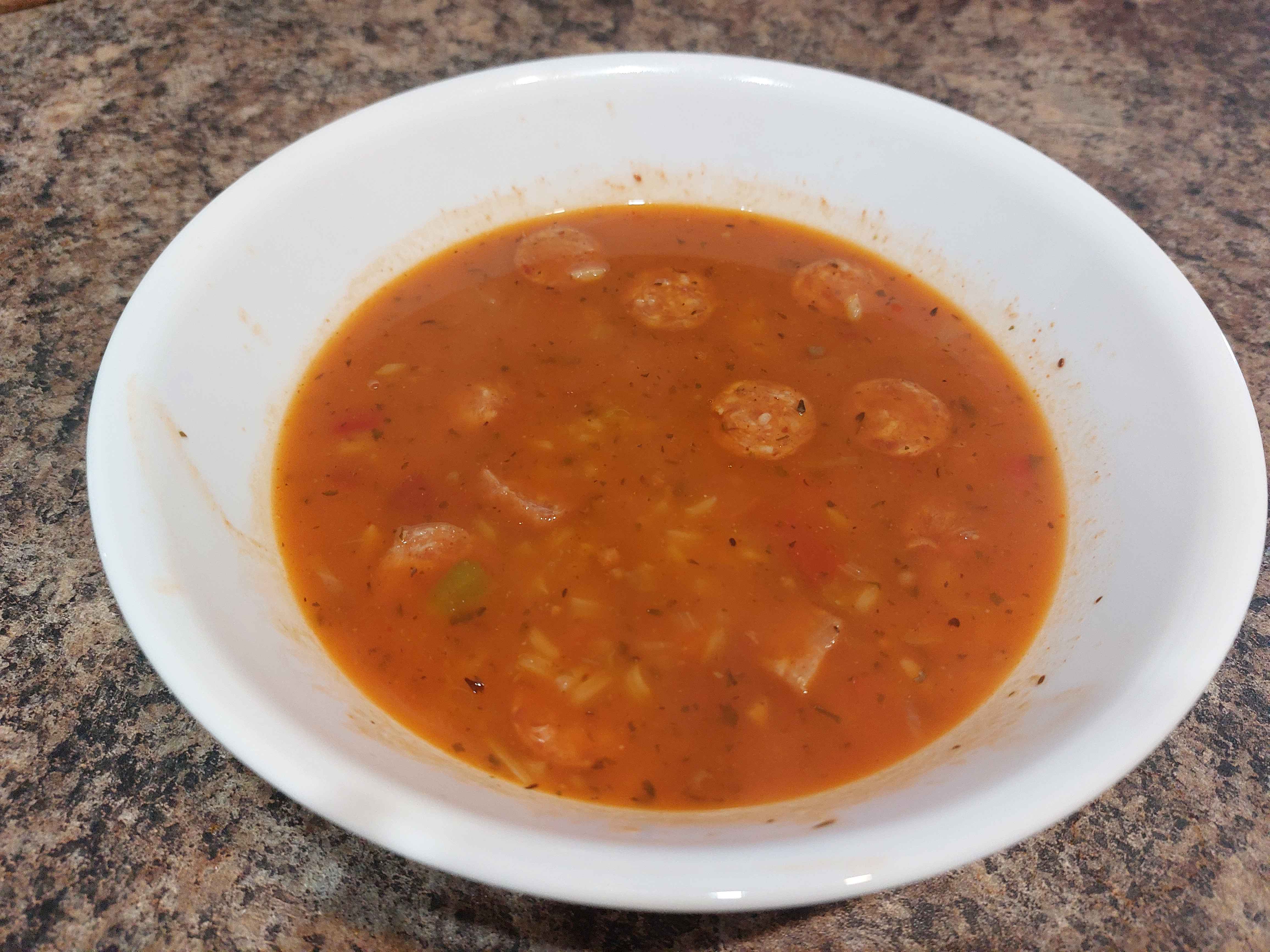 A finished bowl of orange Chicken Gumbo with brown rice, red sausage, and green celery ready for consumption in a white bowl on a brown and black countertop