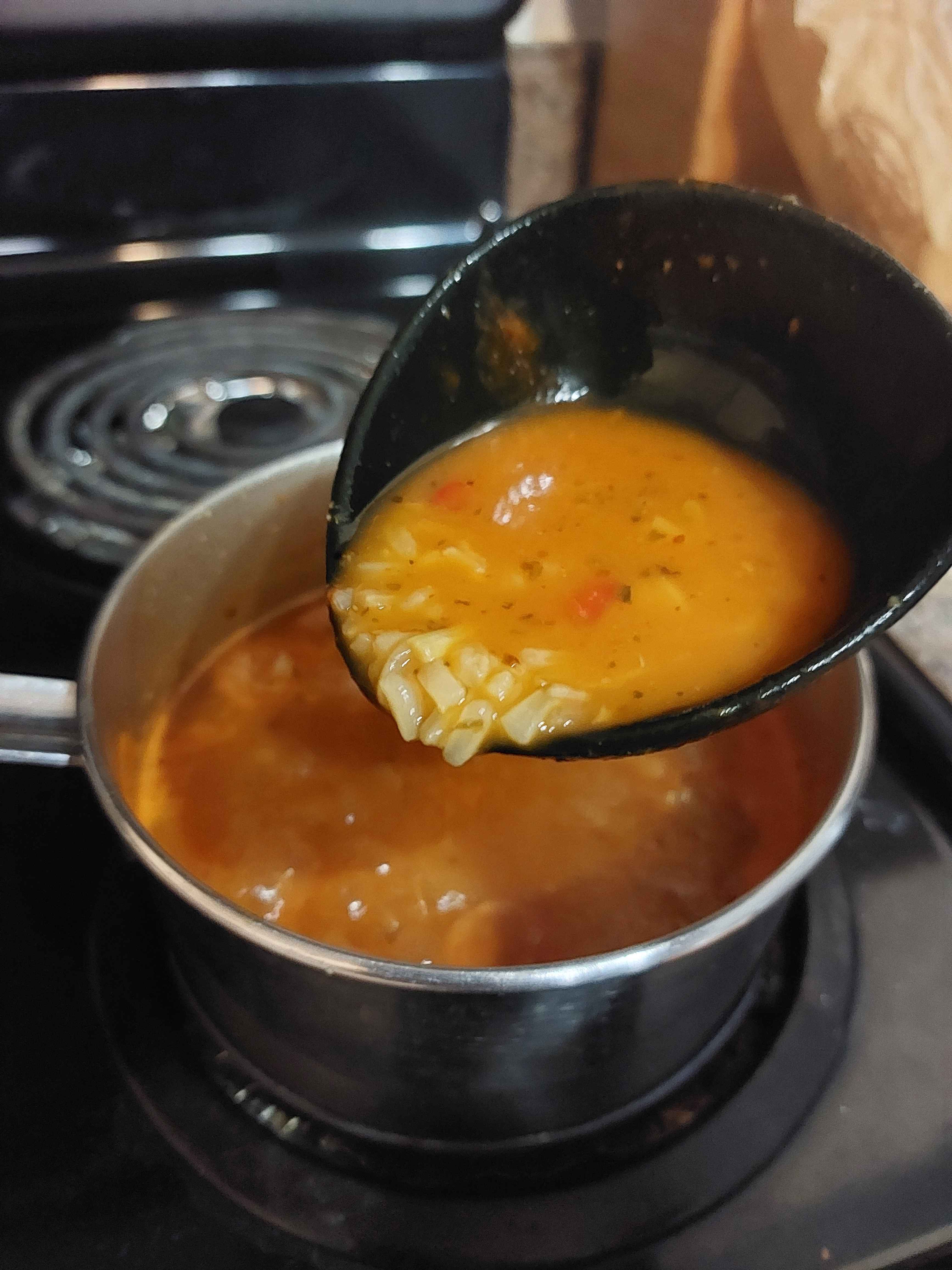 Orange Chicken Gumbo in a silver pot on a black stove boiling while being scooped out using a black ladle including brown rice and red sausage